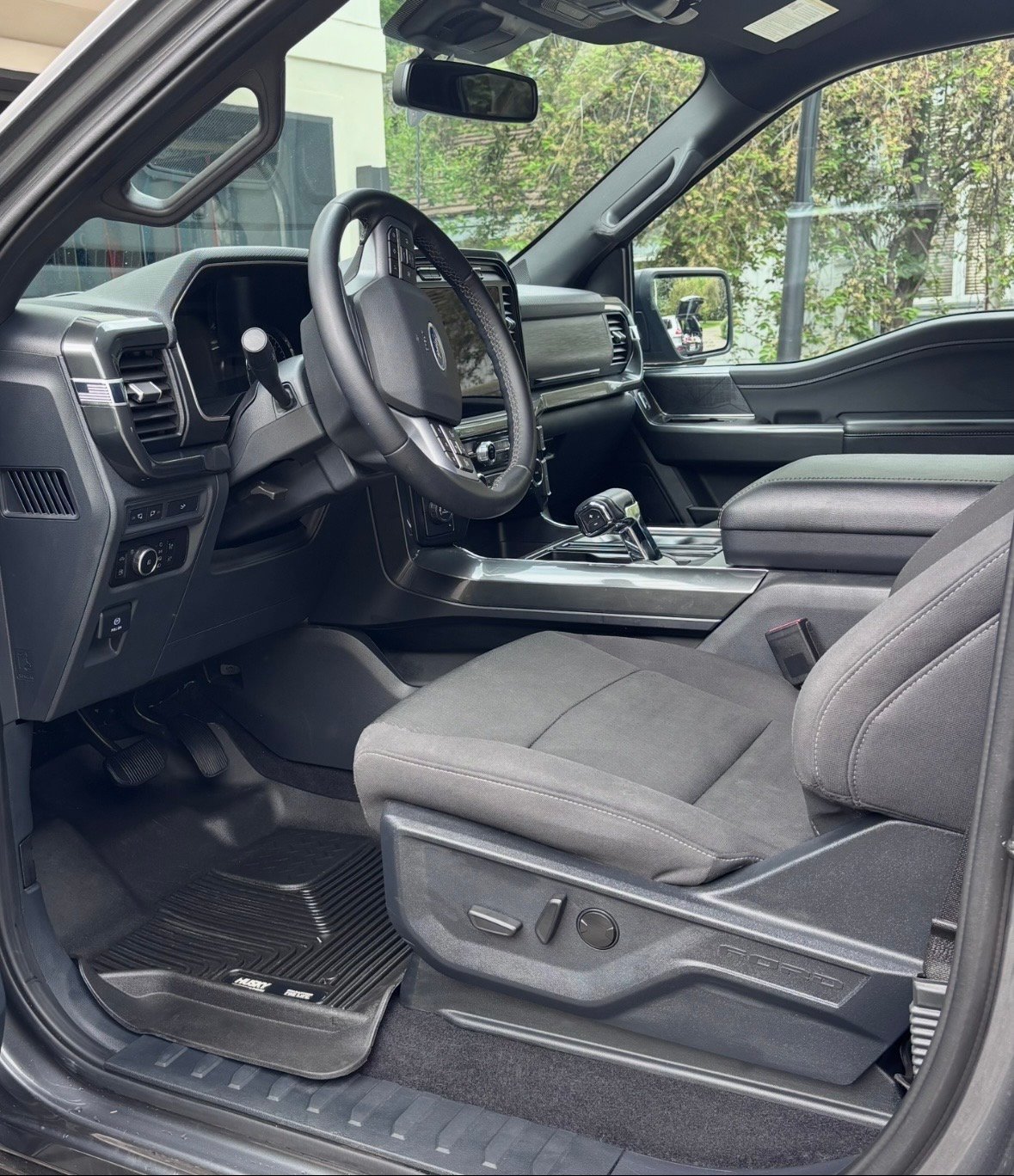Interior of modern truck cabin with steering wheel, dashboard, gray leather seats, and windshield view of trees