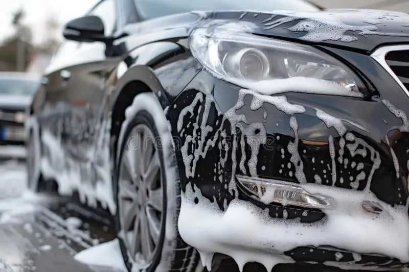 Black sedan covered in soapy foam during outdoor car wash, close-up of front grille and headlight