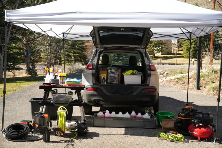 Open car trunk with camping gear and supplies organized on table and ground under white pop-up canopy