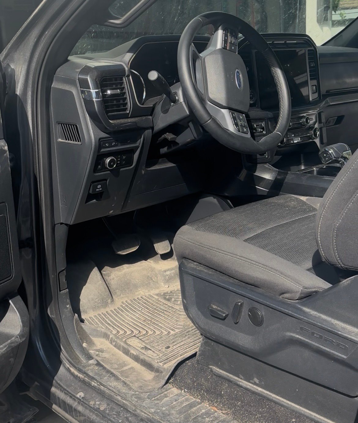Interior view of a vehicle showing steering wheel, dashboard, and front seat with open driver door and visible floor mat